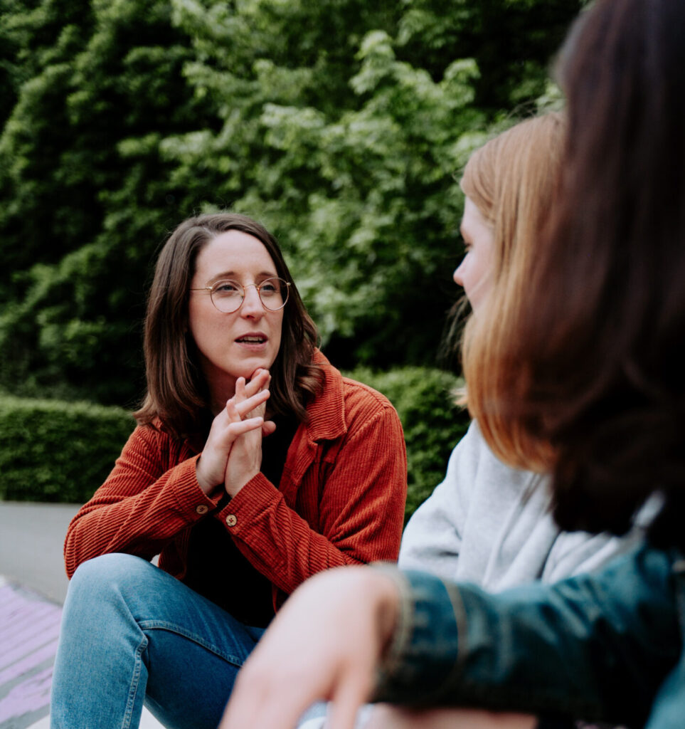 Swantje Schendel sitzt mit zwei jungen Frauen im Park und unterhält sich mit ihnen.